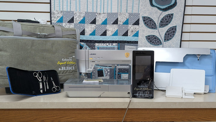 A sewing machine kit displayed on a wooden table, including a gray carrying case, a black tool pouch with scissors and pins, a white and gray sewing machine, a black handheld device, and a blue fabric component, set against a quilted wall display.